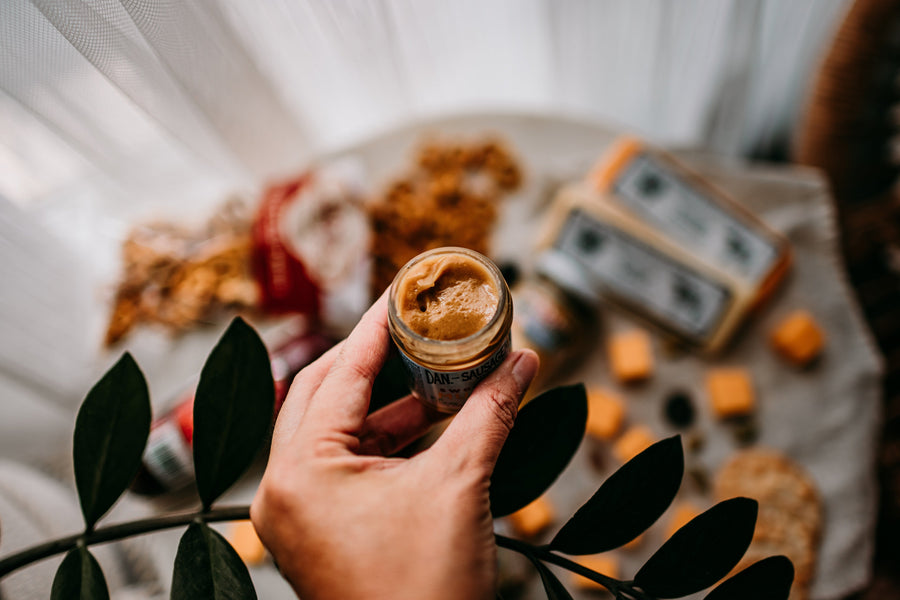 Hand holding a jar of grainy mustard with gift foods in the background; serving suggestion
