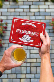 Hands holding Beer & Bourbon Liquor Nuts Men's Gift Tin and glass of beer against brick background.