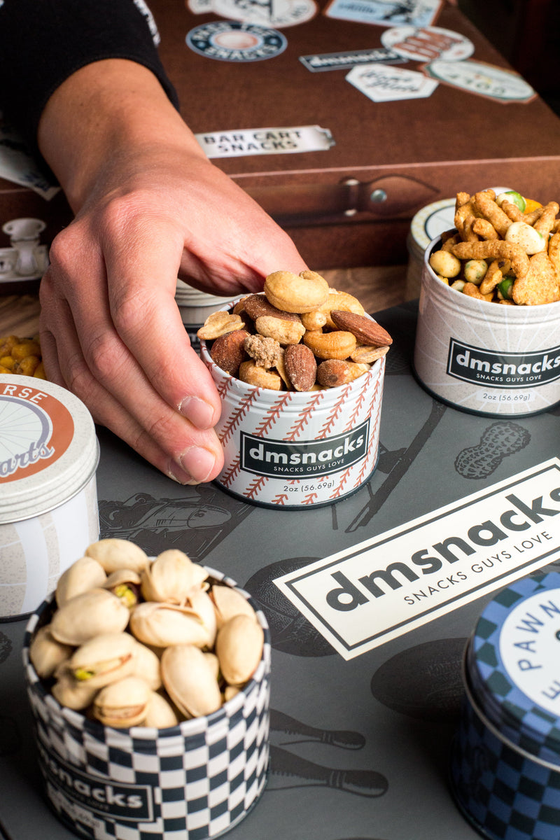 Hand grabbing a tin of assorted nuts from a vintage bar cart filled with various Bar Cart Snacks, including pistachios and trail mix.