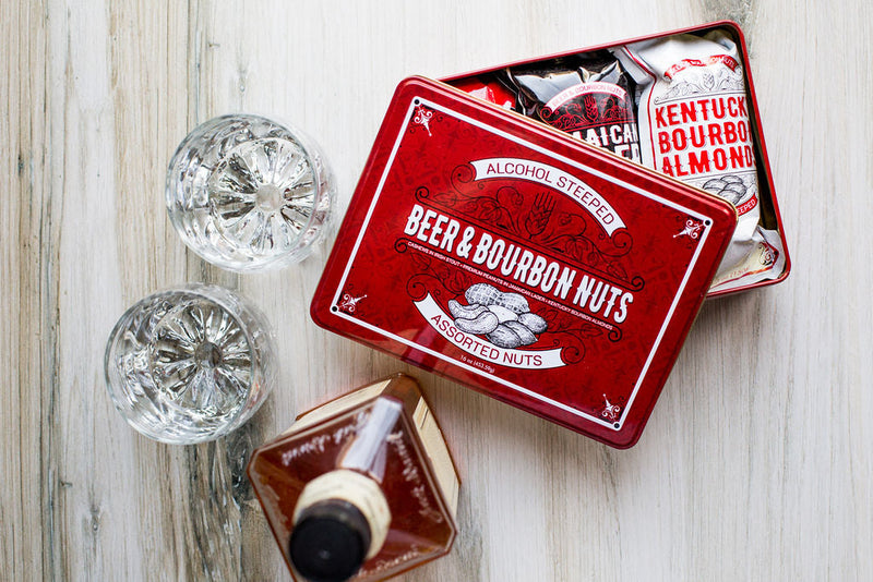 Beer & Bourbon Liquor Nuts Men's Gift Tin with assorted nuts, bourbon bottle, and two whiskey glasses on wooden surface
