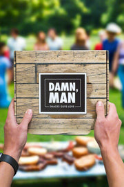 Hands holding a Damn Man Nuts & Beef Box with blurred background of people and picnic table full of snacks.