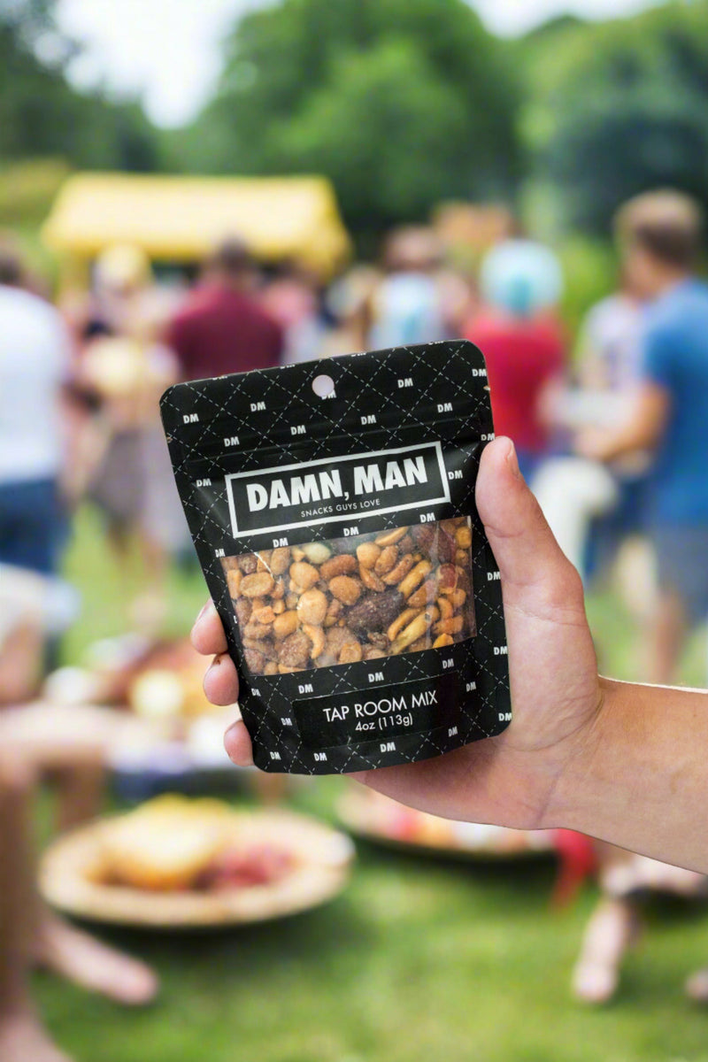 Person holding Damn Man Tap Room Mix nuts snack bag at an outdoor gathering.