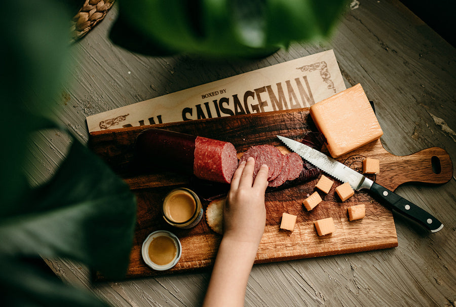 Hands arranging sliced sausage and cheese beside a Dan The Sausageman gift box