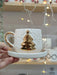 Hand holding a handmade ceramic espresso cup with a gold Christmas tree and star design, paired with a matching saucer.