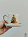 Hand holding a handmade ceramic espresso cup with a gold Christmas tree and star design, paired with a matching saucer.
