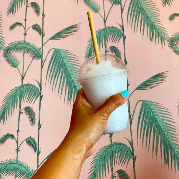 A hand with bright blue nail polish is holding an iced lemonade drink in a clear cup with a dome lid. A bamboo-like straw made from reed stems is inserted through the lid. The background features a pale pink wall with green palm tree designs, giving a tropical and refreshing feel to the scene. The eco-friendly straw and palm backdrop emphasize a playful, sustainable aesthetic.