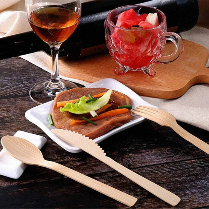 Eco-friendly bamboo cutlery from Holy City Straw Co. displayed on a wooden table next to a square white plate with a grilled steak garnished with a lettuce leaf and carrot strips. A glass of amber-colored drink and a cup of fruit salad complete the cozy dining setting.