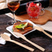 Eco-friendly bamboo cutlery from Holy City Straw Co. displayed on a wooden table next to a square white plate with a grilled steak garnished with a lettuce leaf and carrot strips. A glass of amber-colored drink and a cup of fruit salad complete the cozy dining setting.