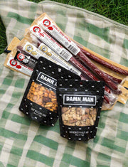 Damn Man Nuts & Beef Box with assorted beef sticks and nut mixes on a green plaid blanket.