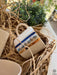 A white ceramic mug with a golden rim, adorned with blue evil eye patterns, partially hidden in a nest of natural straw, resting on a woven placemat, with a blurred background of green plants, presenting a cozy and inviting atmosphere.