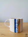 A white ceramic mug with a polished golden handle, detailed with a vertical strip of blue and white evil eye motifs, standing on a wooden surface against a neutral light blue background.