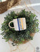 A white ceramic mug with a golden rim and handle, decorated with blue evil eye amulets, nestled in a lush green artificial plant wreath, set on a woven circular placemat with stray raffia strands, on a white surface.
