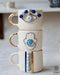 A stack of three white ceramic mugs with golden handles, each featuring a different blue evil eye design; the top with an eye, the middle with a hamsa hand, and the bottom with a stripe of multiple evil eyes, against a soft white background with light blue accents.