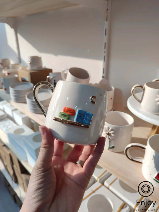 A hand presents a white ceramic mug with a shiny gold handle and a quaint books-and-crescent-moon motif, set in a sunny pottery studio.