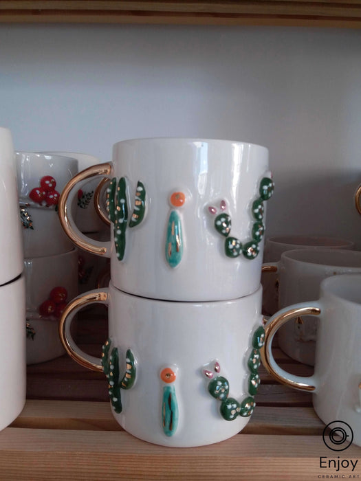 Stacked white mugs with gold handles, adorned with 3D cactus designs and tiny colorful blooms on a wooden shelf.