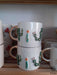Stacked white mugs with gold handles, adorned with 3D cactus designs and tiny colorful blooms on a wooden shelf.