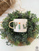  a white ceramic mug with a glossy gold handle, nestled in lush greenery. The mug is embellished with a raised, hand-painted cactus design in vibrant colors, offering a touch of nature's beauty to the setting.