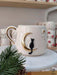 Handmade ceramic mug featuring a black cat sitting on a golden crescent moon, adding a mystical touch to your beverage experience, displayed on a marble stand with holiday accents.