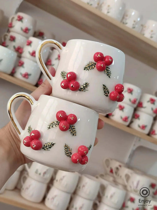 A pair of handmade ceramic mugs featuring festive red berries and intricate green leaves, complete with a golden handle, adding a cheerful touch to any holiday table setting.