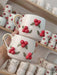 A pair of handmade ceramic mugs featuring festive red berries and intricate green leaves, complete with a golden handle, adding a cheerful touch to any holiday table setting.