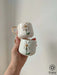 A hand holds two stackable white mugs with gold handles and 3D daisy designs, captured in soft light against a neutral background.