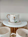 A white ceramic mug featuring a 3D daisy with a gold center and handle, resting on a matching saucer atop a golden pedestal.