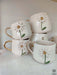 A collection of white ceramic mugs adorned with 3D daisy designs and gold handles, stacked neatly.