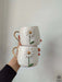 Two white ceramic mugs featuring a 3D daisy design with gold handles, stacked together against a plain background.