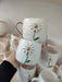 Two white ceramic mugs with daisy designs and gold handles, stacked and displayed among other similar mugs in the background.