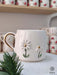 Handmade ceramic mug featuring two delicate daisies with golden centers, accented by a gold handle, displayed on a marble stand with festive greenery.
