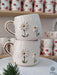Handmade ceramic mugs featuring two daisies with gold centers and a golden handle, stacked on a marble stand with festive greenery.