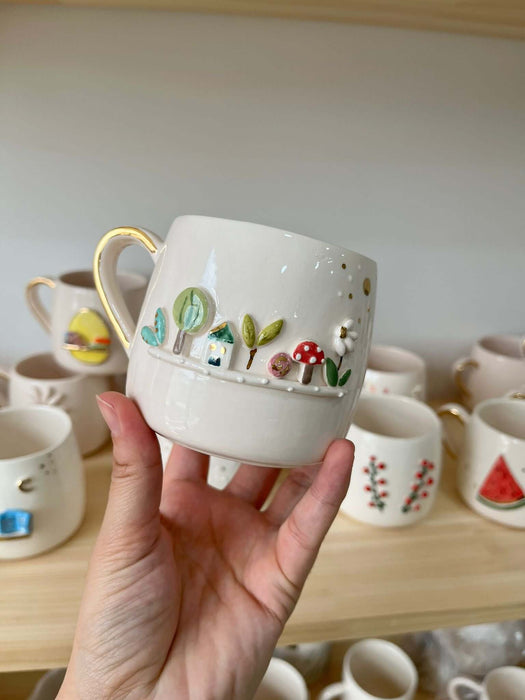 A handcrafted white ceramic mug with a 3D garden scene featuring trees, mushrooms, and a house, held in front of a display shelf.