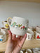 A handcrafted white ceramic mug with a 3D garden scene featuring trees, mushrooms, and a house, held in front of a display shelf.