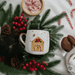 A festive ceramic mug featuring a cozy gingerbread house design, surrounded by pine branches, red berries, and candy canes, creating a warm holiday scene.