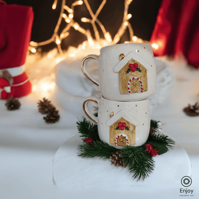 Two stacked ceramic mugs featuring a festive gingerbread house design, surrounded by pine branches and pinecones, glowing against a backdrop of twinkling holiday lights.