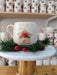 Handmade ceramic mug featuring a gingerbread house design with festive red berries, displayed on a marble stand with pine and pinecone accents, bringing a cozy holiday feel.