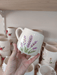 Handmade ceramic mug with lavender design and gold handle, held in front of a ceramic shelf.