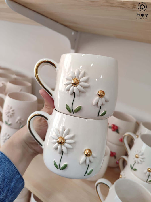 Two handmade ceramic mugs with charming daisy flower designs, featuring golden centers and handles. Each mug is crafted with two daisies, showcasing intricate petal detailing. The mugs are displayed against a shelf filled with other beautiful ceramic pieces.