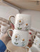 Two handmade ceramic mugs with charming daisy flower designs, featuring golden centers and handles. Each mug is crafted with two daisies, showcasing intricate petal detailing. The mugs are displayed against a shelf filled with other beautiful ceramic pieces.