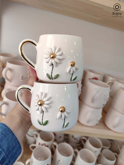 Two ceramic mugs with daisy designs, gold centers, and handles, are displayed in front of a shelf of ceramics.