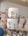 Two ceramic mugs with daisy designs, gold centers, and handles, are displayed in front of a shelf of ceramics.