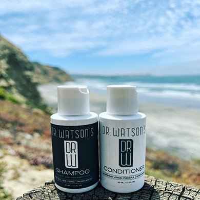 Travel-size Dr. Watson's shampoo and conditioner bottles beside the ocean