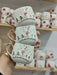A trio of festive snowman espresso cups with gold handles, showcasing intricate holiday designs, held in front of a mug display shelf.
