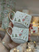 Pair of snowman-themed espresso cups with gold handles, held against a festive backdrop of fairy lights and ceramic mug shelves.