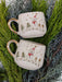 Set of two snowman-themed espresso cups with gold accents, placed on lush greenery for a festive and elegant holiday presentation.