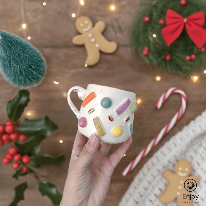 Hand holding a ceramic mug adorned with colorful sprinkles against a festive backdrop of holiday decor, including gingerbread, a candy cane, and a Christmas wreath.