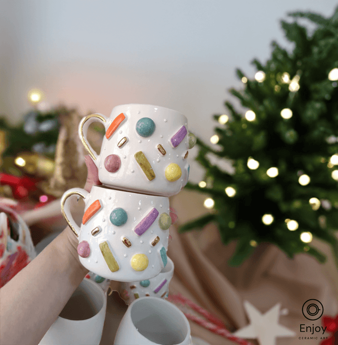 Hand holding two stacked ceramic mugs with colorful sprinkles design, set against a festive Christmas backdrop.