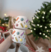 Hand holding two stacked ceramic mugs with colorful sprinkles design, set against a festive Christmas backdrop.