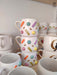 Three handmade mugs stacked on a wooden shelf, featuring vibrant pastel sprinkles in orange, yellow, green, purple, and pink with gold accents, offering a playful and cheerful aesthetic.