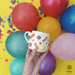 Hand holding a festive ceramic mug with colorful sprinkles, set against a backdrop of vibrant balloons and confetti.